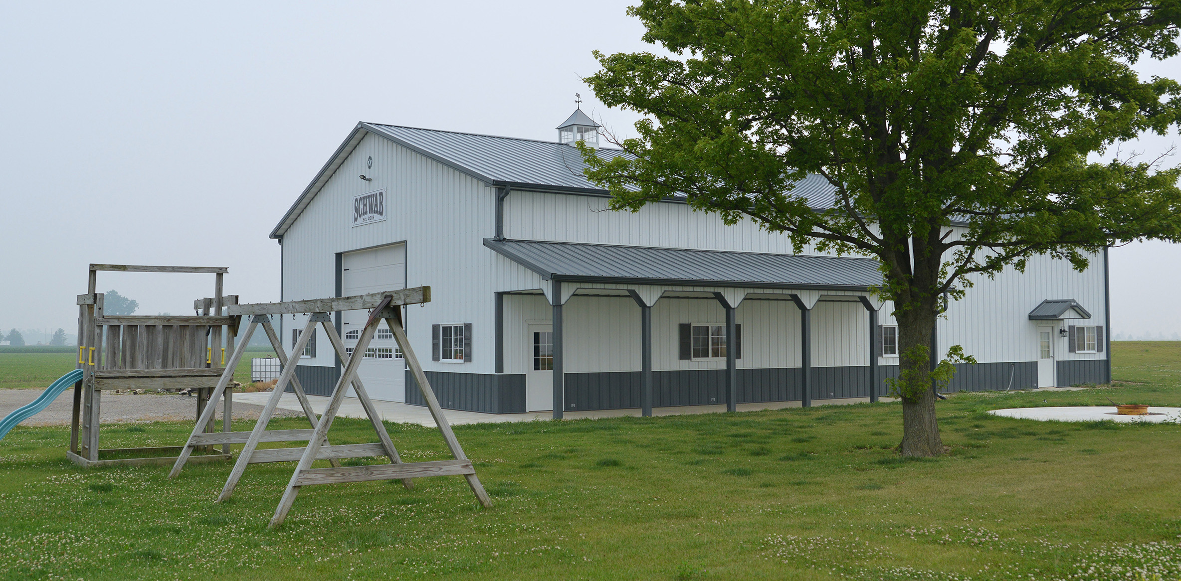 Gray and white post frame suburban building with a tree and small playground in the yard